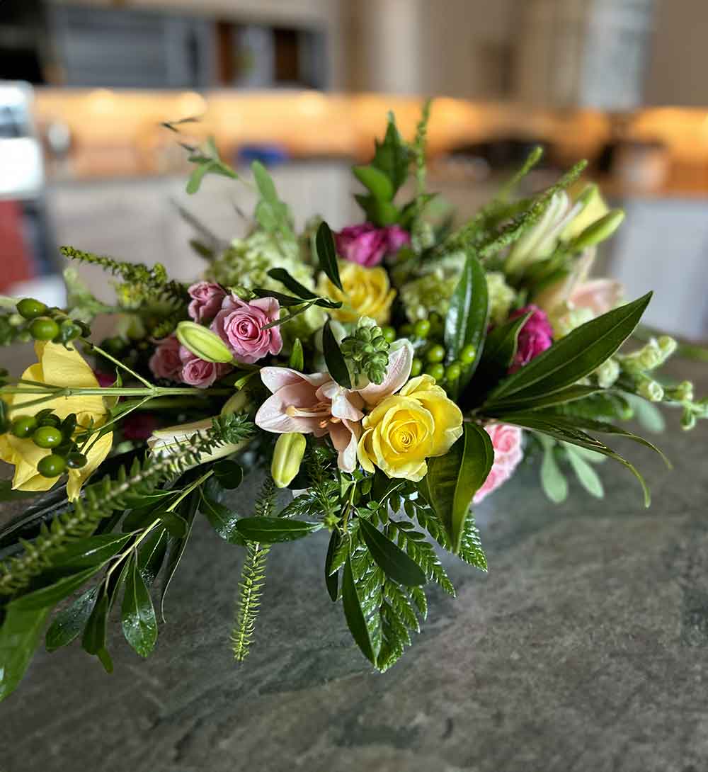 A vibrant "Low Centerpiece" arranged on a table, featuring a mix of yellow roses, pink roses, lilies, and lush greenery in a well-lit indoor setting. The background is softly blurred, drawing attention to the colorful floral arrangement.