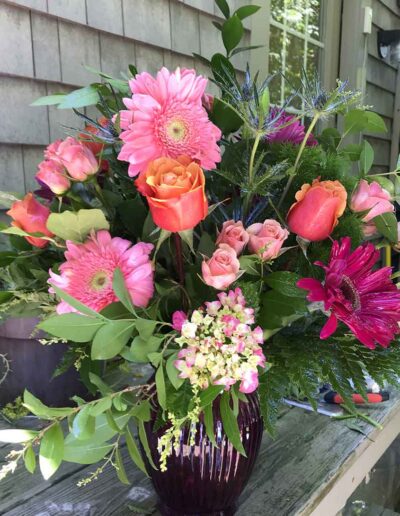 A vibrant bouquet named "Large" in a purple vase, featuring pink gerbera daisies, orange and pink roses, small white flowers, and assorted greenery, is placed on a wooden table outdoors with a shingle-sided building in the background.