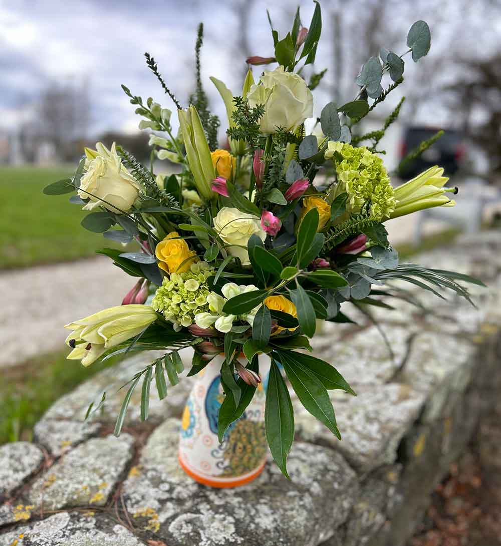 A vibrant bouquet featuring large yellow roses, smaller yellow roses, white blossoms, and lush green foliage. The flowers are artistically arranged in a natural style, creating a fresh and cheerful display. Introducing Medium, the perfect arrangement for any occasion.