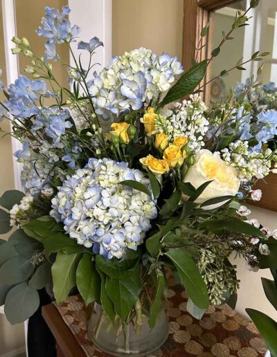 A Large glass vase filled with an arrangement of pastel-colored flowers sits on a table. The bouquet includes white and light blue hydrangeas, small yellow roses, white blossoms, and various green leaves and stems. A mirror and part of a staircase are visible in the background.