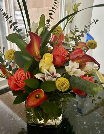 A vibrant floral arrangement from the Medium collection in a glass vase sits on a dark countertop. This bouquet features an assortment of colorful flowers including red and yellow roses, orange calla lilies, white freesia, yellow billy balls, and lush greenery. A white window provides a serene backdrop.