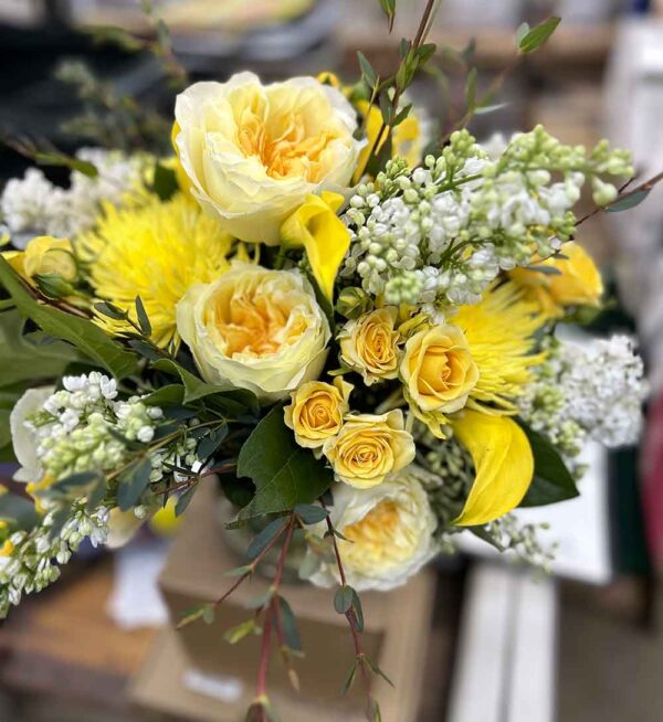 A vibrant bouquet featuring large yellow roses, smaller yellow roses, white blossoms, and lush green foliage. The flowers are artistically arranged in a natural style, creating a fresh and cheerful display. Introducing Medium, the perfect arrangement for any occasion.