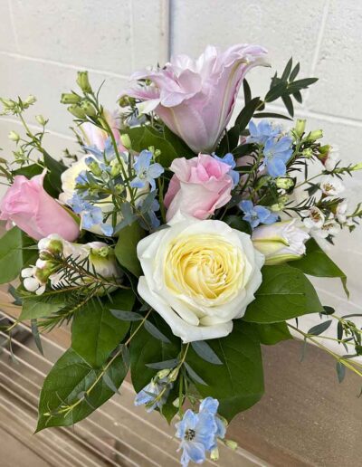 The Medium bouquet showcases an elegant arrangement of white, pink, and purple blooms, including exquisite roses, complemented by lush green foliage and delicate blue flowers. The flowers are artfully arranged in a neat cluster and set against a wooden surface with a white tile background.