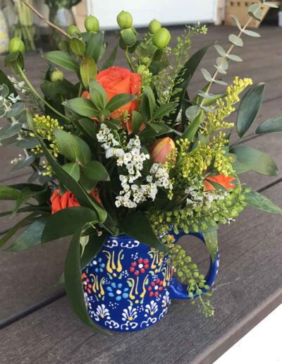 A vibrant floral arrangement featuring orange roses, white flowers, and various green foliage in a colorful blue and white patterned ceramic mug named "Small," placed on a wooden surface.