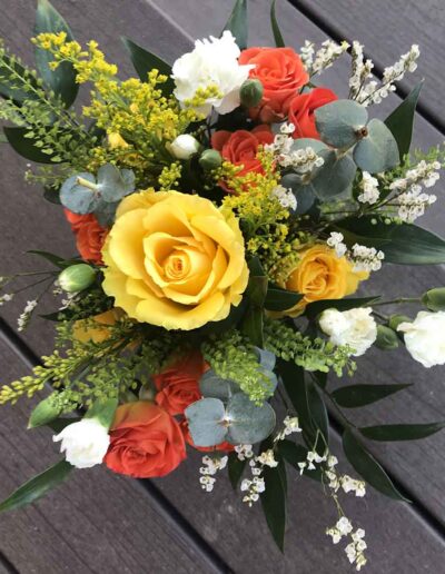 A "Small" bouquet of flowers featuring prominently yellow and orange roses, along with white buds and assorted green foliage, displayed against a dark wooden background. The arrangement has a fresh and vibrant appearance.