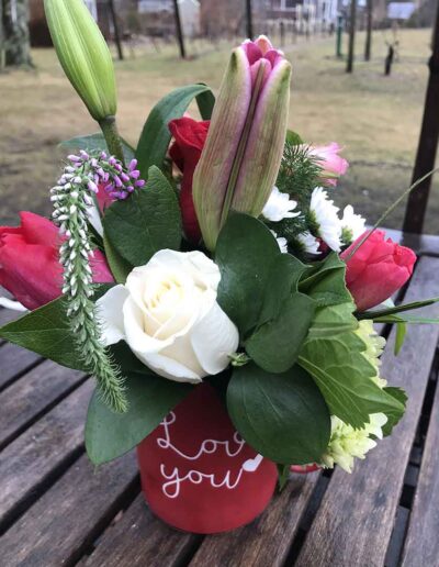 A bouquet of flowers in a red vase with the words "Love you" written in white. The bouquet includes a variety of flowers such as white roses, red tulips, and unopened lilies, arranged with green leafy foliage. This exquisite arrangement from Small is beautifully displayed on a wooden table outdoors.