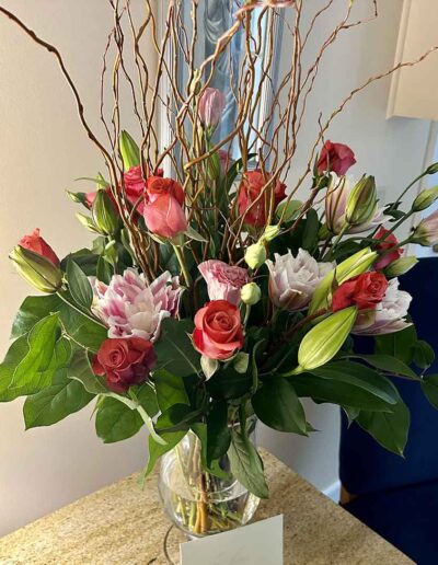 A bouquet of various flowers, including roses, lilies, and greenery, is arranged in a clear glass vase placed on a wooden table. The bouquet is accented with curly willow branches, and there is a white card in front of the vase labeled "Extra Large." A blue chair is visible in the background.