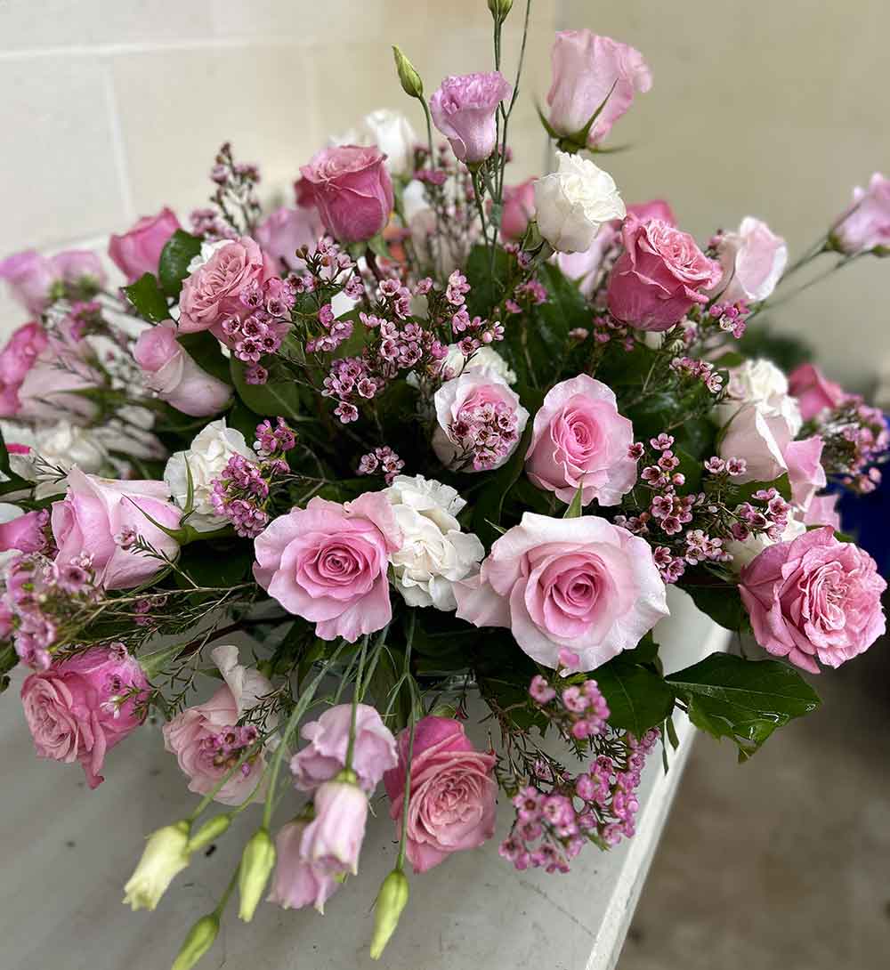A captivating floral display in our "Extra Large" arrangement, showcasing a diverse mix of pink roses, white carnations, and petite purple blossoms, all beautifully enhanced with rich green foliage. This exquisite bouquet sits elegantly on a white table set against a simple backdrop.