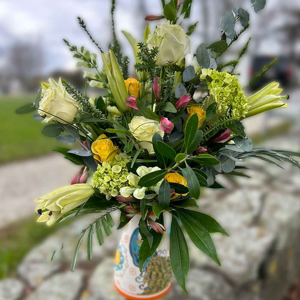 A vibrant bouquet featuring large yellow roses, smaller yellow roses, white blossoms, and lush green foliage. The flowers are artistically arranged in a natural style, creating a fresh and cheerful display. Introducing Medium, the perfect arrangement for any occasion.