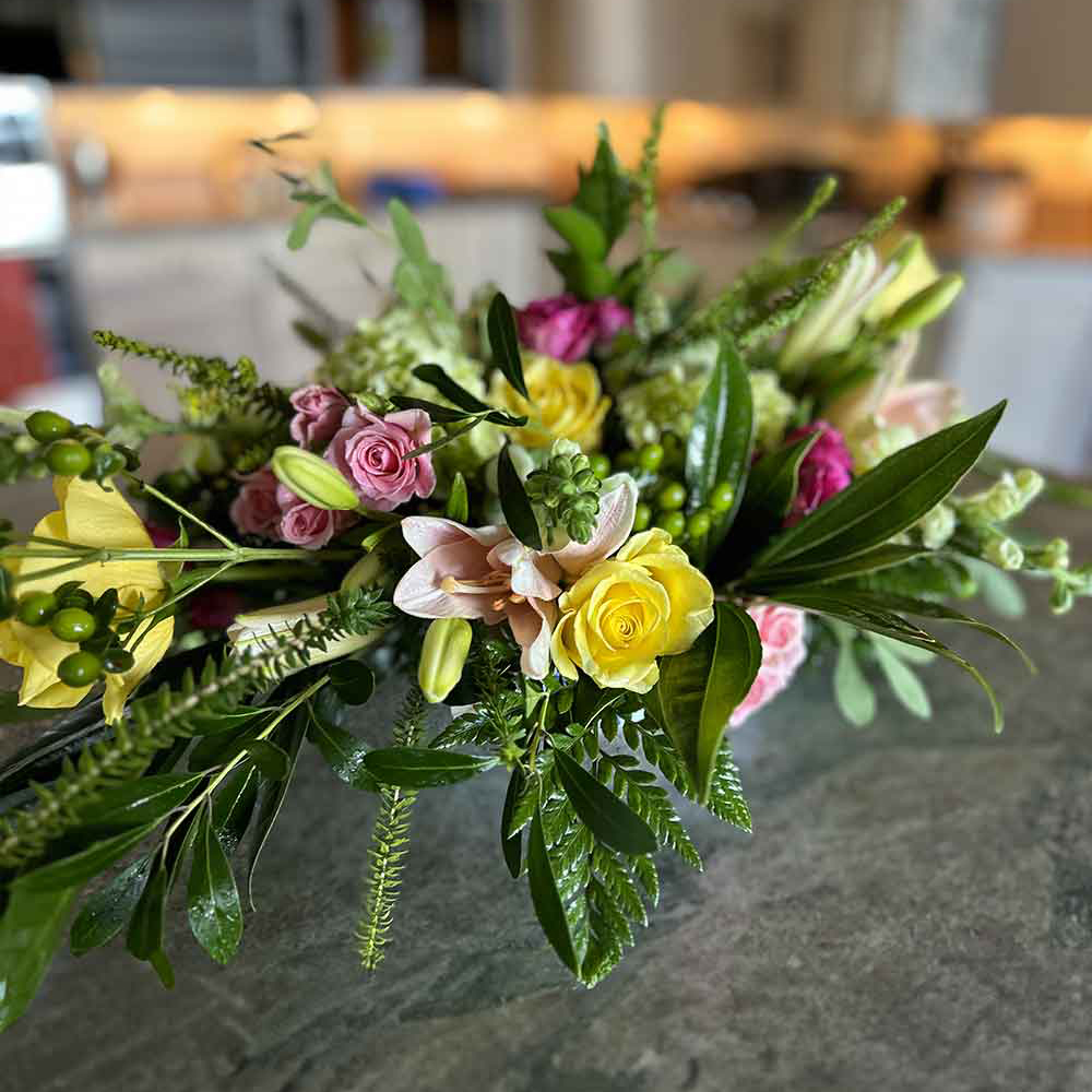 A vibrant "Low Centerpiece" arranged on a table, featuring a mix of yellow roses, pink roses, lilies, and lush greenery in a well-lit indoor setting. The background is softly blurred, drawing attention to the colorful floral arrangement.