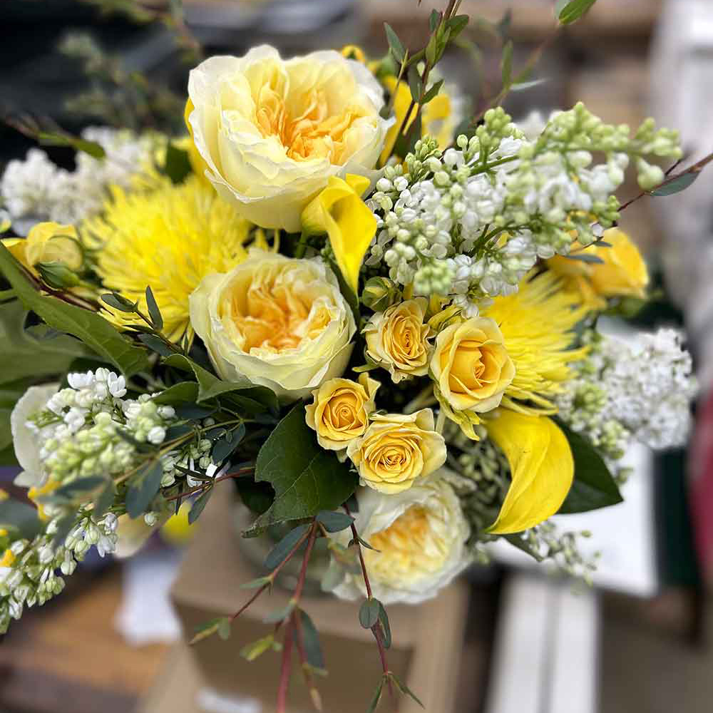 A vibrant bouquet featuring large yellow roses, smaller yellow roses, white blossoms, and lush green foliage. The flowers are artistically arranged in a natural style, creating a fresh and cheerful display. Introducing Medium, the perfect arrangement for any occasion.