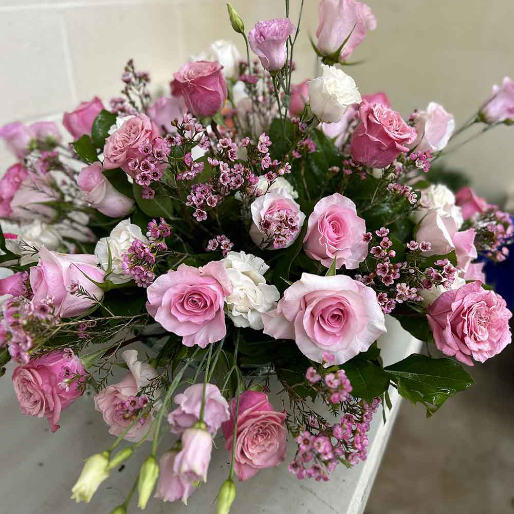 A captivating floral display in our "Extra Large" arrangement, showcasing a diverse mix of pink roses, white carnations, and petite purple blossoms, all beautifully enhanced with rich green foliage. This exquisite bouquet sits elegantly on a white table set against a simple backdrop.